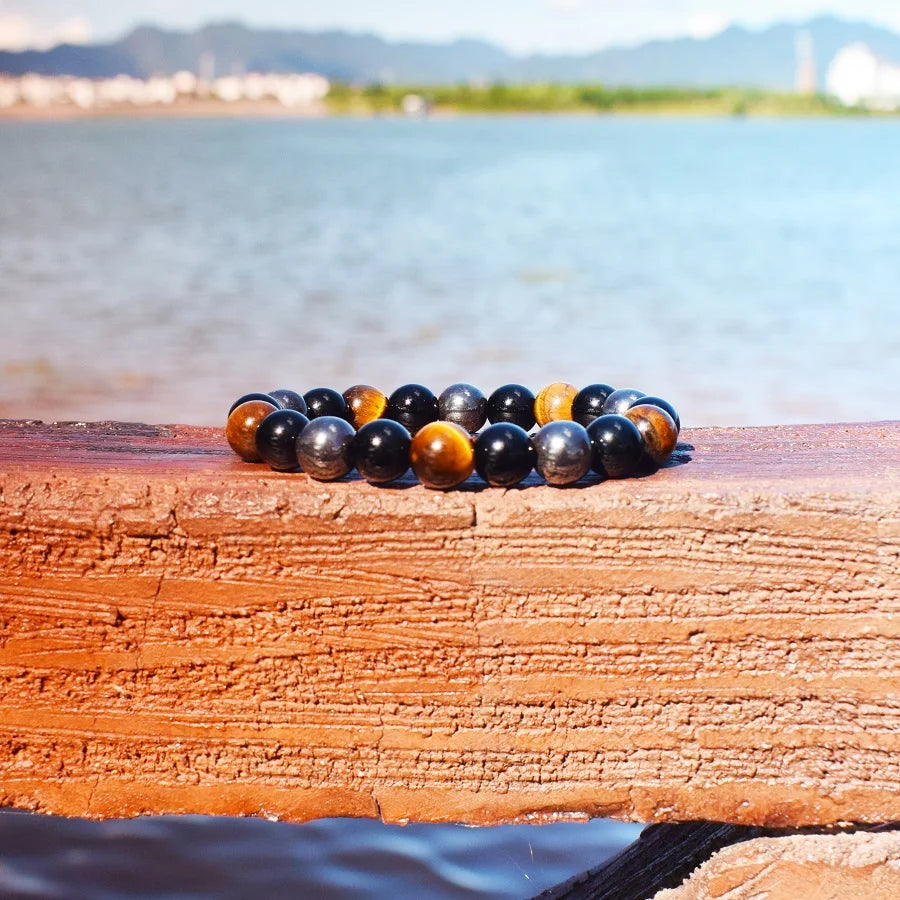 Bracelet magnetique pour maigrir homme avec perles d'oeil de tigre et hematite sur bois flotte face a l'ocean